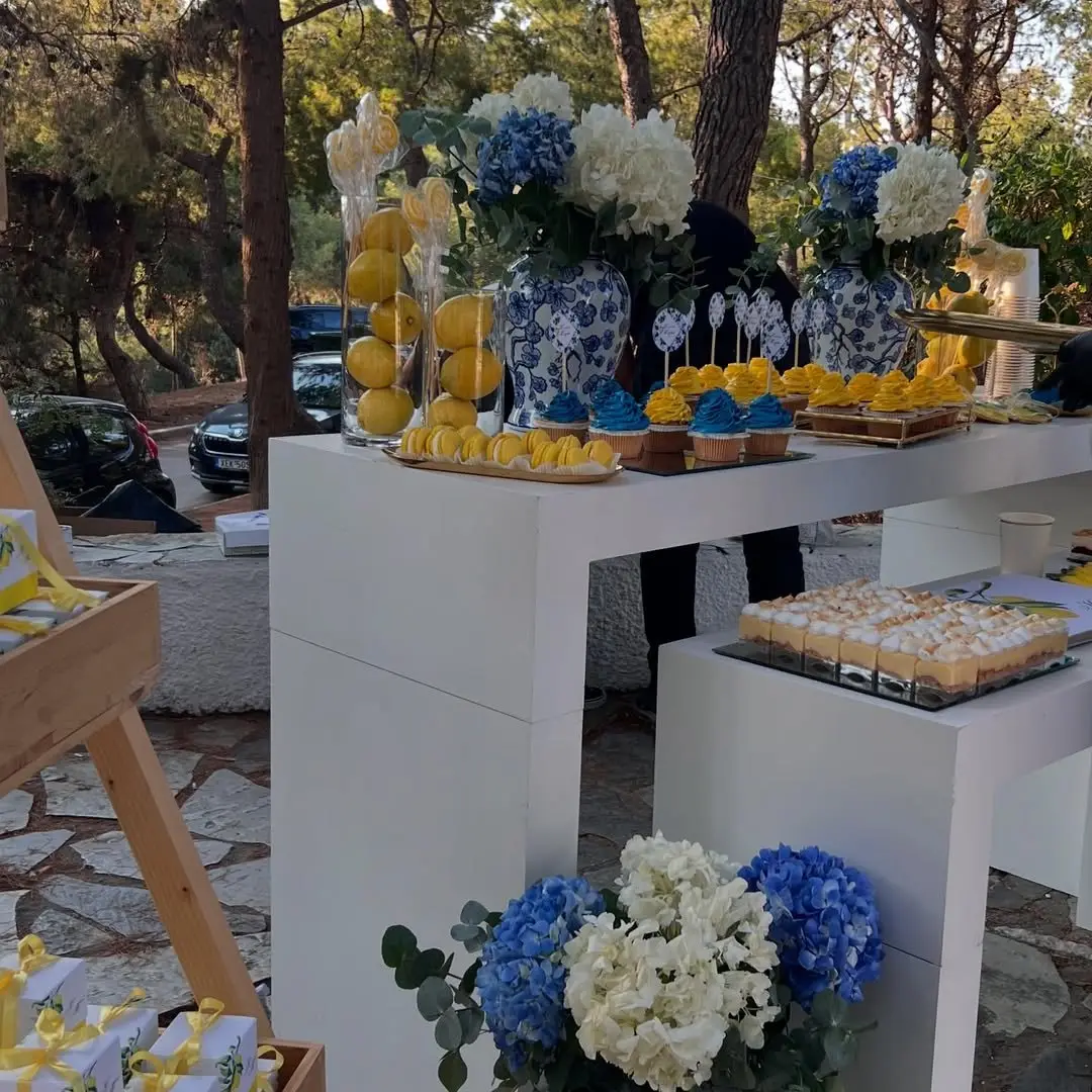 Candy bar βάπτισης με θέμα λεμόνι, μπλε-λευκά βάζα και λουλούδια σε κήπο